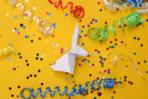 Origami rabbit with colored streamers and confetti on a yellow background Easter celebration