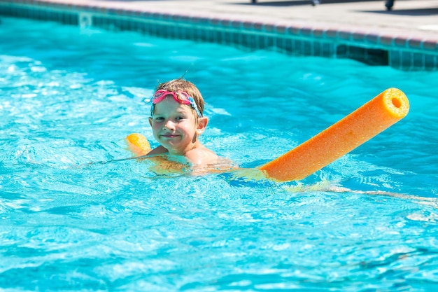 Oudoor summer activity Concept of fun health and vacation Happy smiling boy five years old in swim glasses swim in the pool with noodle in hot summer day