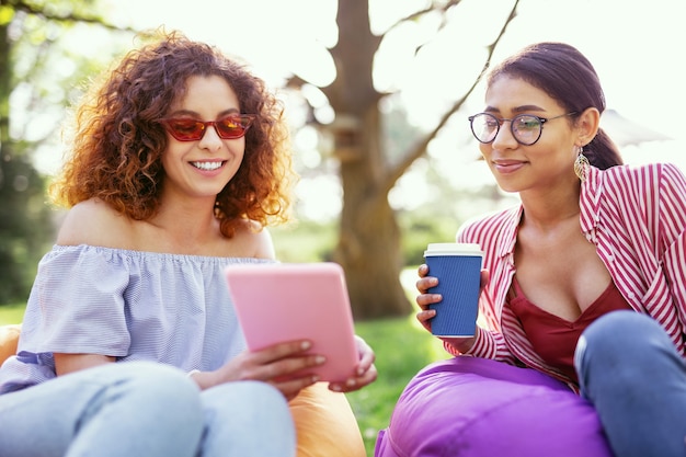 Our friendship. Delighted curly-haired woman holding a tablet and talking with her friend