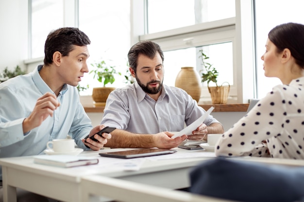 Our startup. Good-looking serious bearded man holding a sheet of paper and talking to his partners sitting near him