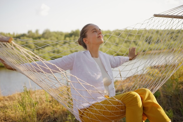 Out door shot of cheerful female sitting on hammock near the river looking up with charming smile enjoying her rest near the water wearing white shirt and yellow trousers