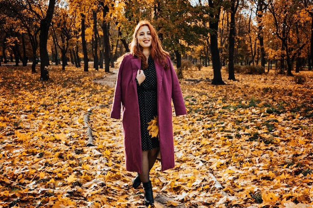 Outdoor autumn portrait of happy smiling plus size red hair woman in coat walking in fall park