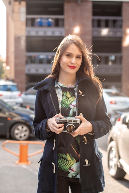 Outdoor autumn smiling lifestyle portrait of pretty young woman, having fun in the city with camera, travel photo of photographer. Making pictures in hipster style.