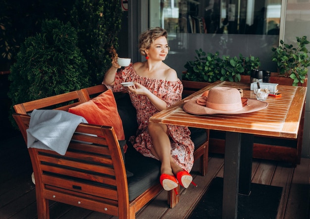 Outdoor fashion portrait of a stunning woman sitting in a cafe I drink coffee and read an old book a woman in a dress and a hat