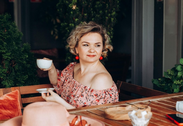 Outdoor fashion portrait of a stunning woman sitting in a cafe I drink coffee and read an old book a woman in a dress and a hat