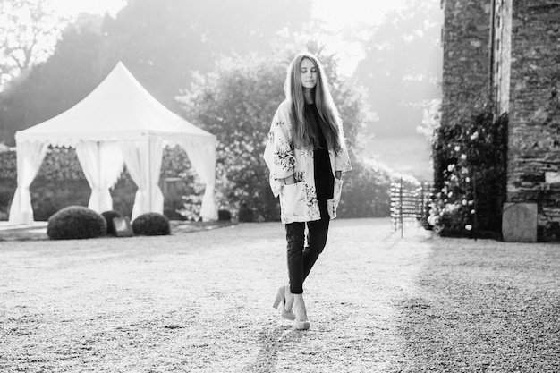 Outdoor horizontal portrait of pleasant looking fashionable woman on high heeled shoes looks pensively down poses at camera stand against wonderful landmarks and white tent Beauty and nature