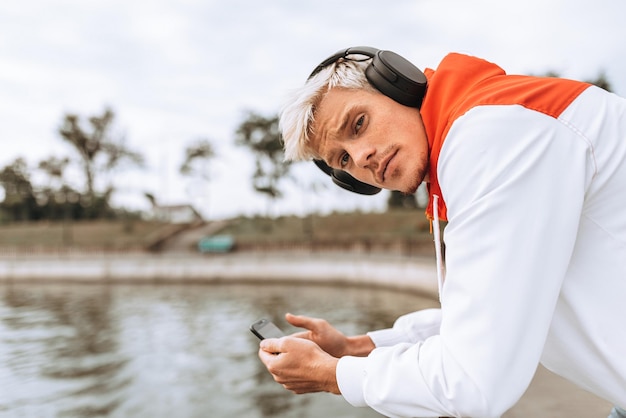 Outdoor image of young man enjoying weather in city park waiting for his girlfriend texting message while listening to his favorite music from wireless headphone using smart phone in the city park