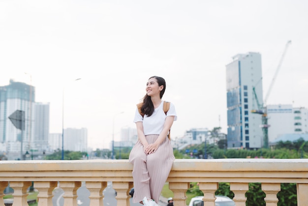 Outdoor lifestyle close up portrait of happy young woman in stylish casual outfit sitting on bridge on the street. Pretty hipster girl having fun and enjoying holidays.