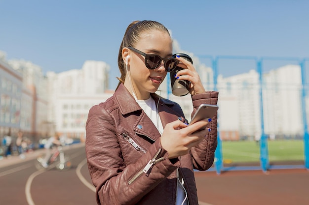 Outdoor photo of attractive student girl in sunglasses with snowwhite smile long hairstyle and wearing jacket chilling in city in sunlight with smartphone