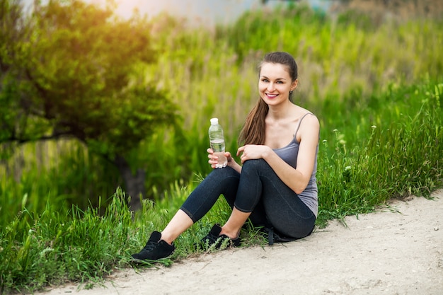 Outdoor photo of pretty young woman wearing sportive clothes and drinking water sitting beside a river