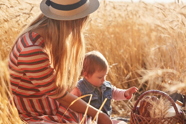 Outdoor picture of young fair haired mother sitting with her little daughter at wheat field, having small picnic together, mum looking after kid, going to eat and drink outside, spending good time.