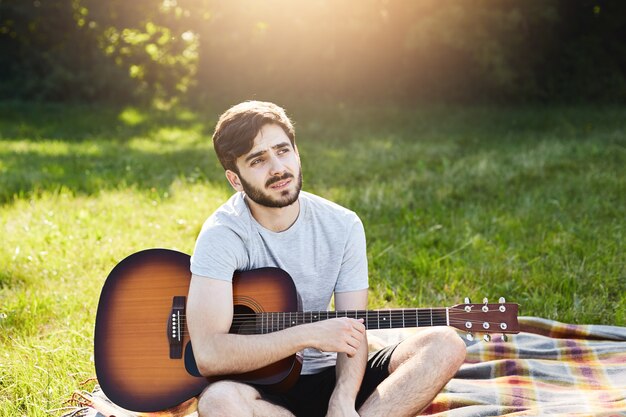 Outdoor portrait of attractive young man with beard dressed casually while resting on green grass