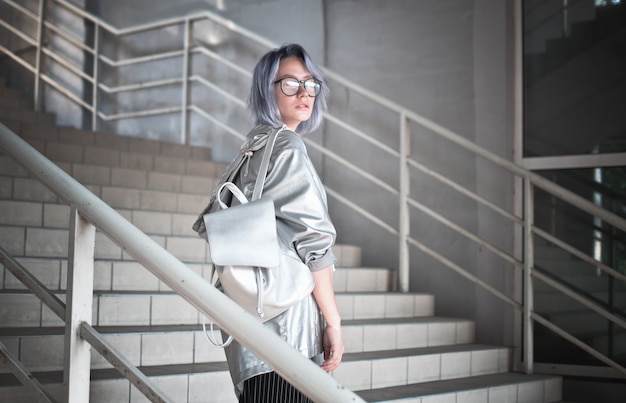 Outdoor portrait of an extraordinary student girl with ashen blue hair dressed in a cloak with a silvery fabric and a backpack on her shoulders.