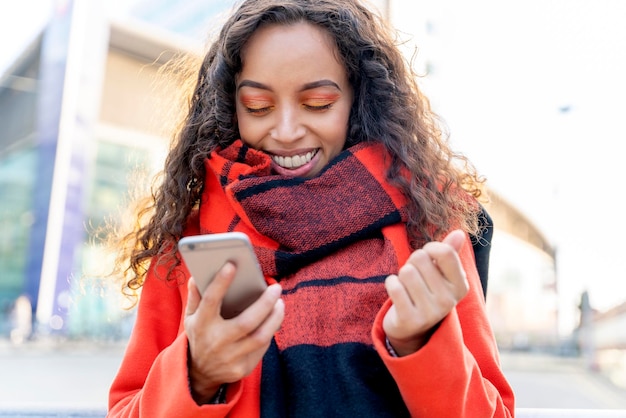 Outdoor portrait of a happy woman in orange coat and scarf using a mobile phone and laughing