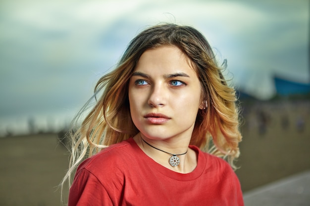 Outdoor portrait of serious white girl about 21 years old with blond hair and red shirt.