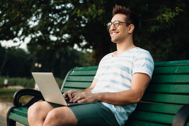 Outdoor shoot of young programmer working on a bench. Side view portrait of a young student sitting with a laptop on her legs smiling.