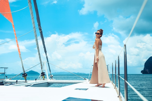 Outdoor shot of adorable young woman in beige dress standing on edge of yacht