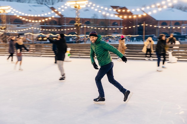 Photo outdoor shot of attractive man with beard wears warm winter clothes practices going skating on ice skate ring decorated with lights has glad expression enjoys his favourite action