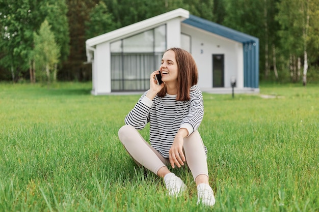 Outdoor shot of attractive positive woman talking with cell phone sitting on grass on yard with big house on background looking smiling away copy space