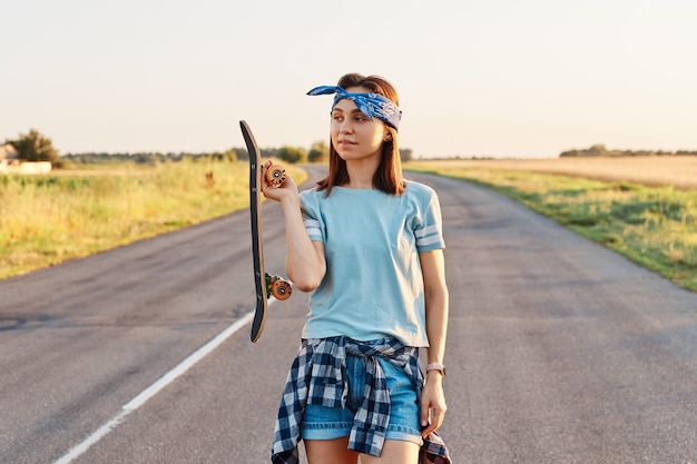 Outdoor shot of beautiful dark haired woman wearing hair band, t shirt and short, holding longboard in hands and looking away with thoughtful expression.