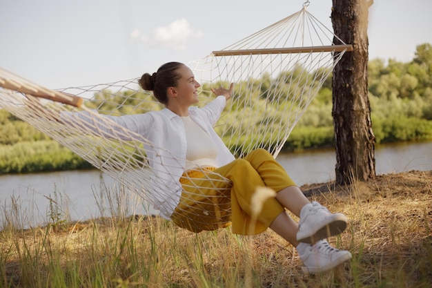 Outdoor shot of Caucasian young adult woman sitting on hammock by the water looking away resting spreads hands laughing happily wearing white shirt and yellow trousers