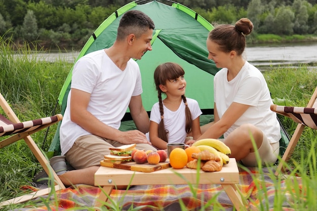 Outdoor shot of happy family sitting near tent on the ground having picnic together enjoying delicious fruit and sandwiches talking and spending happy weekend