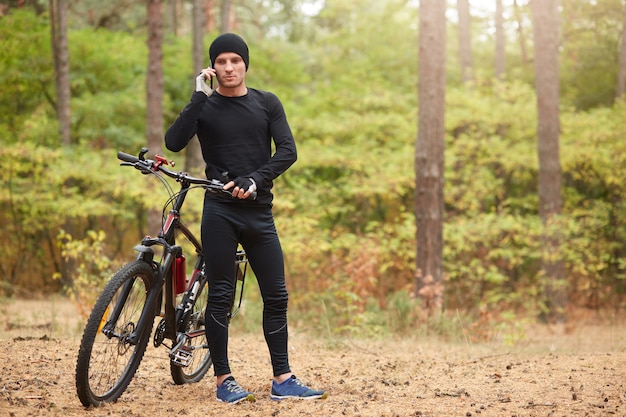 Outdoor shot of man wearing black track suit and sneakers, standing near his mountain bike and speaks phone with beautiful forest on backgraund, spending time in active way. Healthy lifestyle concept.