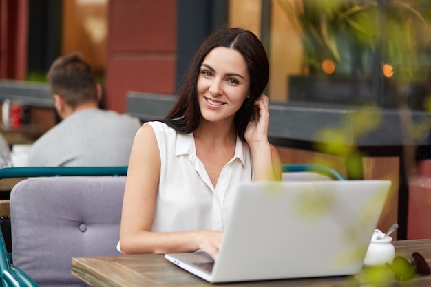 Outdoor shot of pleasant looking brunette young woman with charming smile, spends free time at cozy restaurant, works on laptop computer, searches internet and messages with friends, uses wi fi