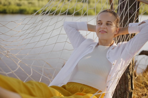 Outdoor shot of relaxed satisfied Caucasian woman wearing white shirt sitting in hammock on the bank of the river raised arms and relaxing near the water