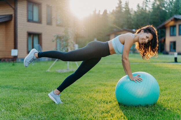 Outdoor shot of slim active sporty woman does fitness exercises with fitball, dressed in active wear, has cheerful expression, poses during sunny day on green grass, being in good physical shape