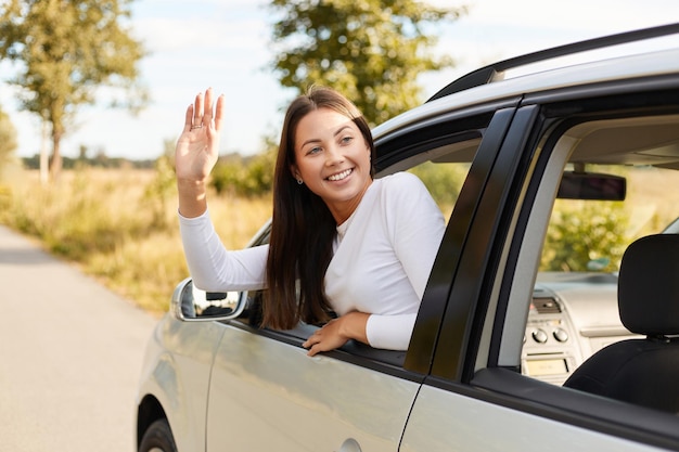 Outdoor shot of smiling happy Caucasian young adult dark haired woman driver waving her hand behind car steering wheel looking back with positive expression saying hello to somebody