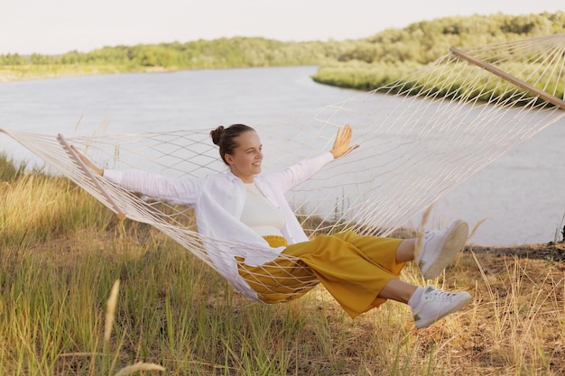 Outdoor shot of smiling satisfied female wearing white shirt sitting on hammock on the bank of the river spreads hands looking away enjoying beautiful nature