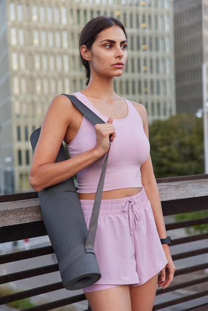 Outdoor shot of sporty woman dressed in sportswear carries rolled rubber karemat going to have fitness training focused into distance rests after training focused into distance. Healthy lifestyle