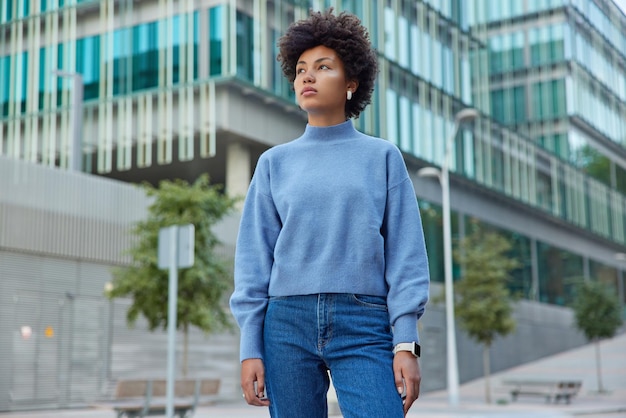 Outdoor shot of thoughtful curly haired woman strolls in modern city concentrated into distance wears casual blue jumper and jeans enjoys free time at street poses against blurred background