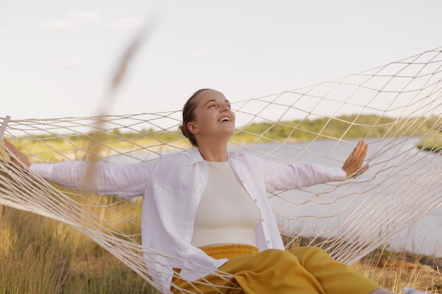 Outdoor shot of young adult Caucasian female wearing white shirt sitting on hammock on the bank of the river looking up at sky and laughing enjoying rest near the water