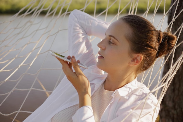 Outdoor shot of young adult woman with bun hairstyle wearing white shirt sitting in hammock on the bank of the river holding mobile phone and recording voice message looking away at beautiful view