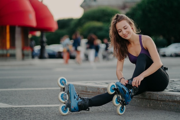 Outdoor shot of young sporty woman in sportswears puts on roller blades poses on road going to ride rollers during sunny day ties shoelaces has good mood Recreation hobby lifestyle concept