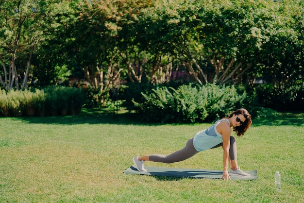 Outdoor sport concept. Healthy fit energetic young woman does fitness exercises on fitness mat dressed in active wear drinks fresh water wears sunglasses poses on green grass. Morning workout.