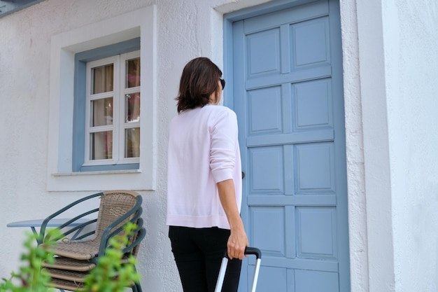 Outdoor woman traveler with yellow suitcase opening closing door of the house