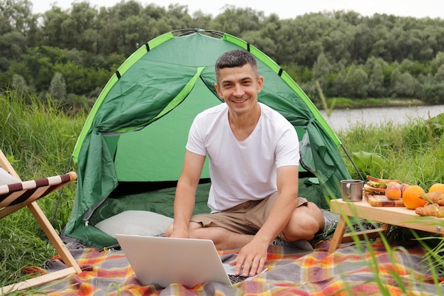 Outdoor young adult man tourist freelancer working online on a laptop outdoors sitting on ground near tent looking at camera with happy satisfied expression