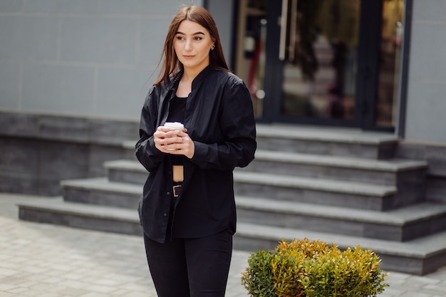 Outdoors lifestyle portrait of stunning brunette girl. Drinking coffee and walking on the city street.