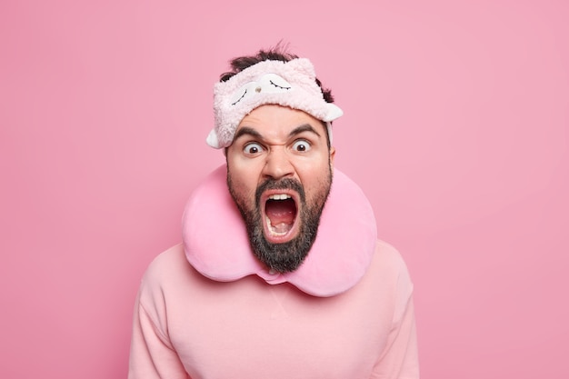 Outraged bearded man screams louldly keeps mouth widely opened expresses negative emotions being angry with someone who interrupts sleep wears blindfold travel pillow around neck 