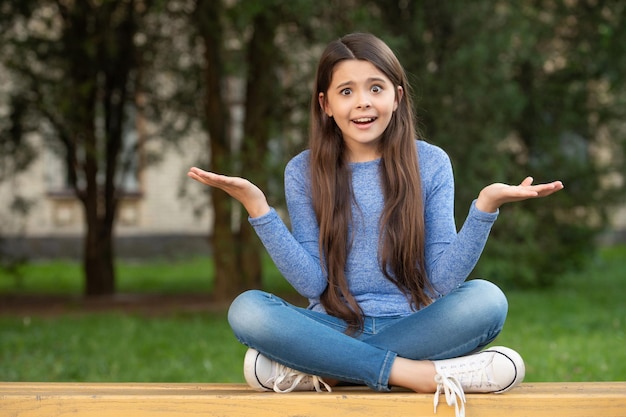 Outraged teenage girl shrugging shoulders sitting legs crossed on bench outdoors outrage