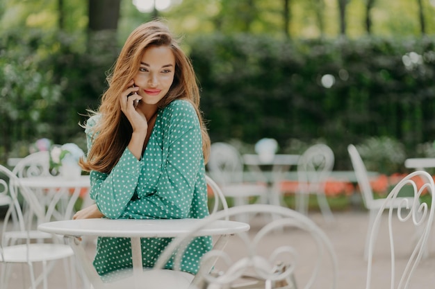 Outside shot of charming young woman with long hair wears polka dot green shirt sits at table in outdoor cafe has pleasant talk via modern smartphone has dreamy expreession People and lifestyle