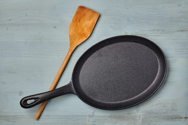 Oval cast iron frying pan with a wooden spatula on a blue wooden table. Template for laying out dishes