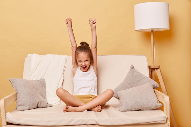 Ovedrjoyed happy positive optimistic little girl wearing casual clothing sitting on sofa at home against beige wall rejoicing her success raised clenched fists