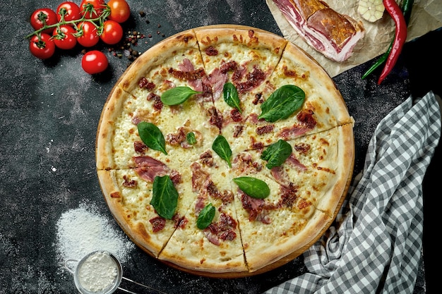 Oven-baked Italian pizza with sauce, cheese, ham and sun-dried tomatoes in a composition with ingredients on a dark table. View from above