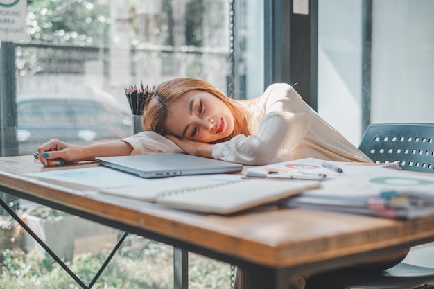 Photo overcome by exhaustion a young woman rests her head on her desk amidst a spread of work materials a moment of stillness in her busy day captured in a lightfilled office space