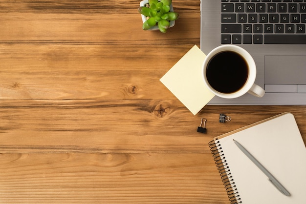 Overhead above close up flat lay view photo of laptop keyboard cup of beverage sticker binder clips spiral copy book with grey pen isolated brown backdrop with empty blank space