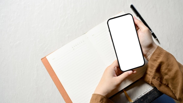 Overhead A female hands holding a smartphone white screen mockup over her white working desk